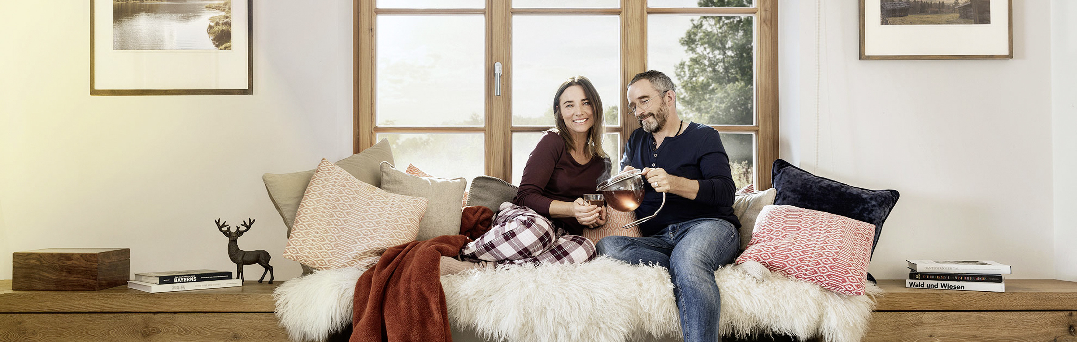 Ein Mann und eine Frau sitzen umgeben von Kissen vor einem Fenster und trinken Tee.