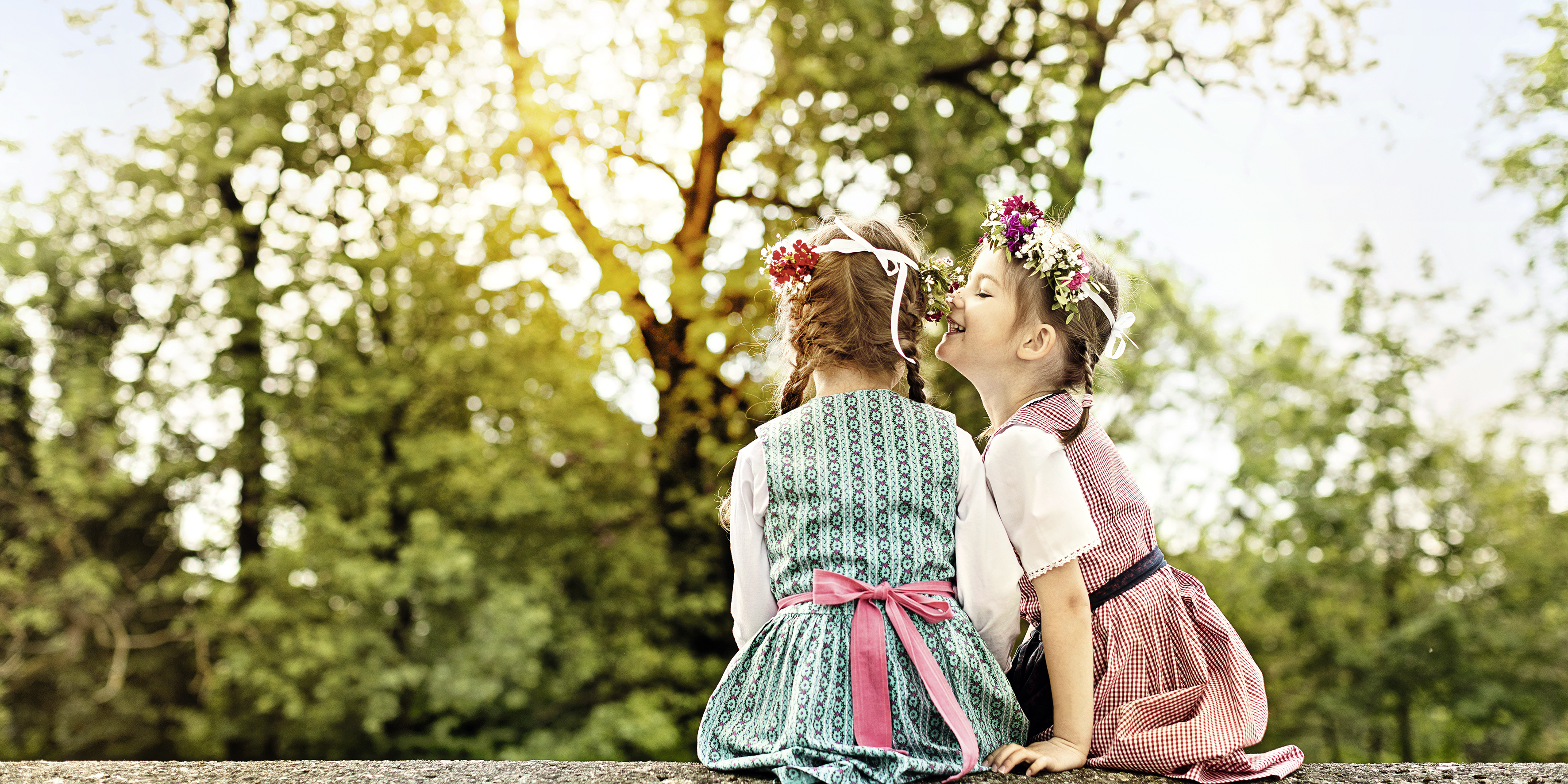 Zwei Mädchen sitzen im Grünen und das eine flüstert dem anderen etwas ins Ohr