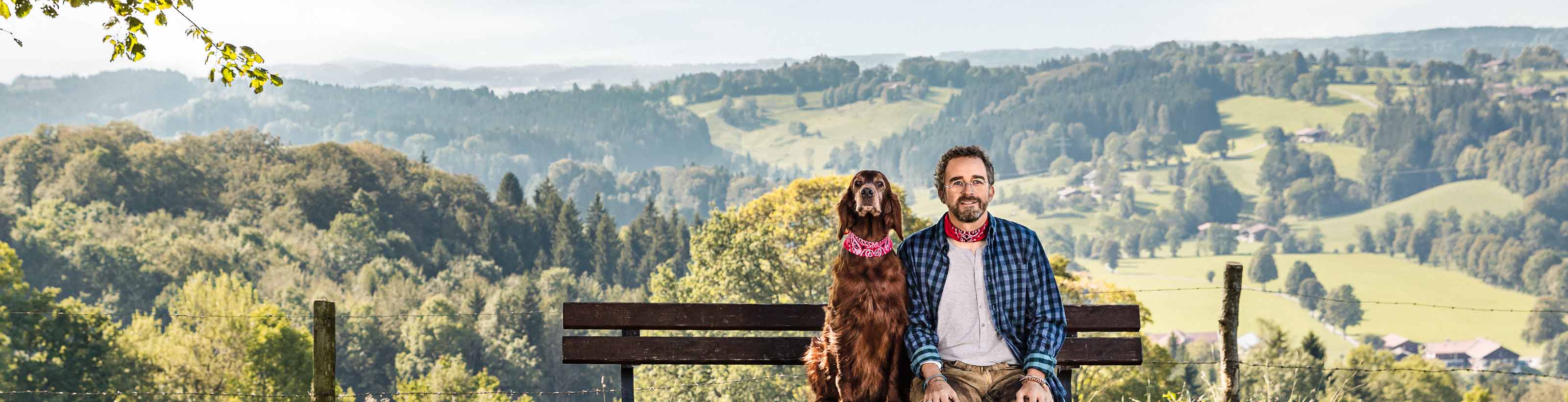 Ein Mann und ein Hund mit roten Halstüchern sitzen auf einer Bank in einer hügeligen Landschaft