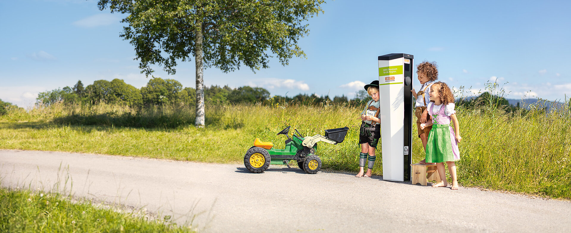 Drei Kinder in Tracht stehen mit einem Trettraktor an einer ESB Ladesäule im Grünen