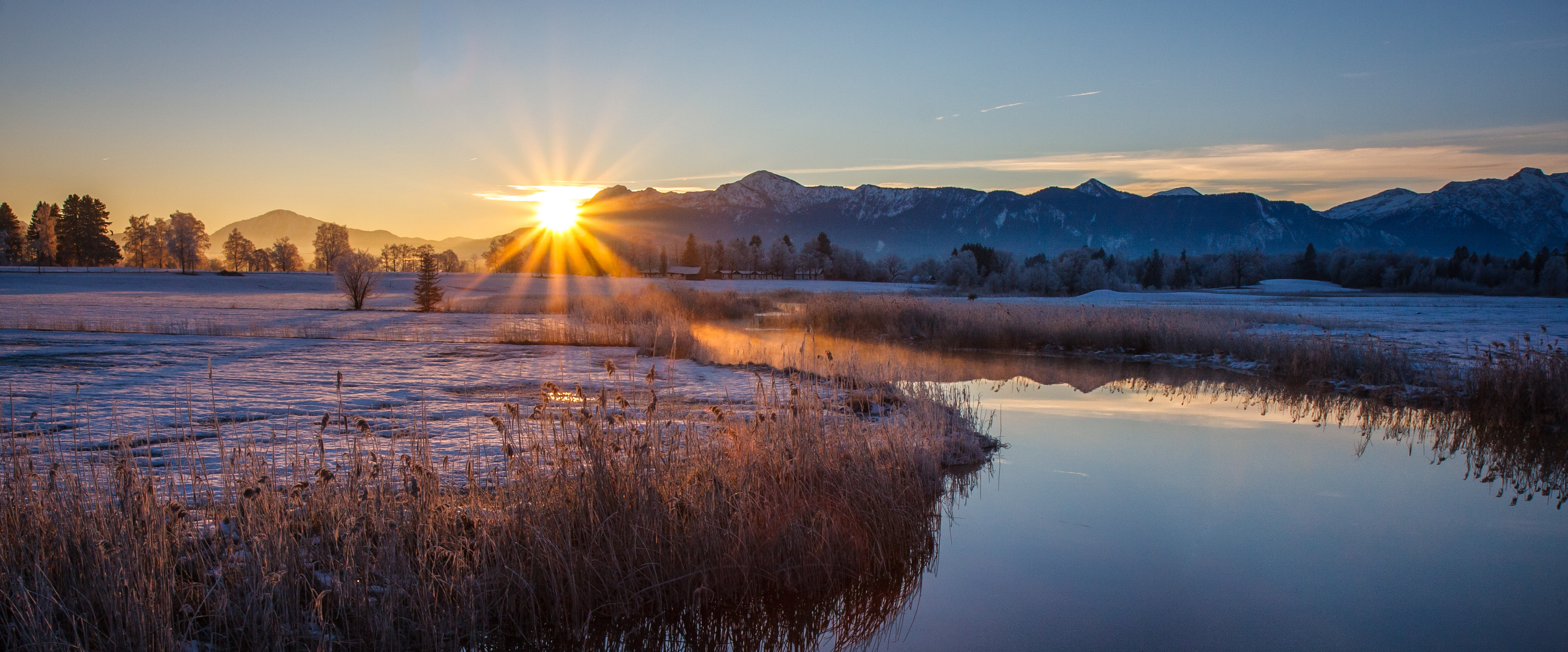 Ein Bach in einer Schneelandschaft mit Bergen und der untergehenden Sonne im Hintergrund