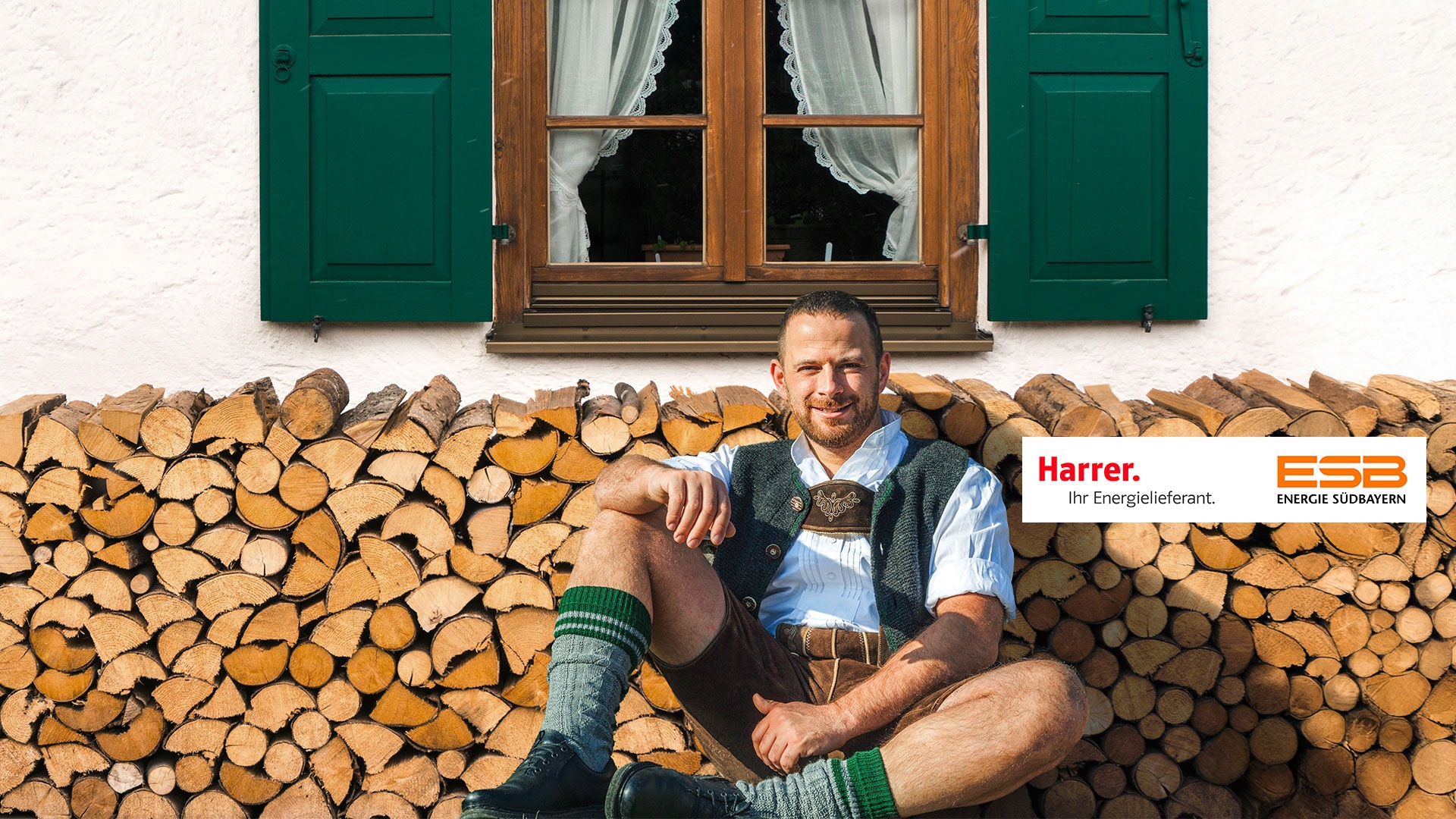 Mann in Lederhose sitzt vor einem Brennholzstapel vor einem Haus.